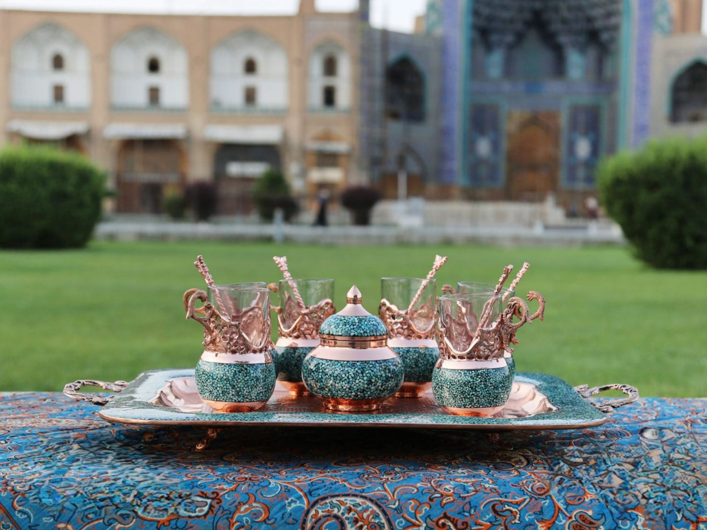 turquoise inlay tea set, Persian tea set in garden, traditional tea set outdoors, handmade tea set with monument background, cultural tea set photography, Isfahan tea set display, copper and turquoise tea ware, Firoozeh Koobi tea set in nature, elegant tea set in Persian garden, luxury tea set with historic backdrop, Iranian handicraft in garden setting