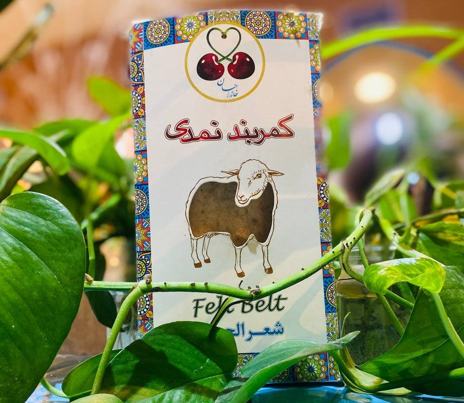 A rectangular product package surrounded by green leaves. The package shows a drawing of a sheep, Persian text reading “kamarband namadi,” and a logo of two cherries forming a heart shape at the top. The design includes colorful decorative borders.