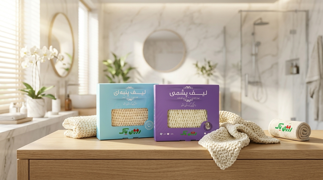Two boxed natural-fiber bath scrub mitts on a wooden counter in a bright bathroom, with knitted beige scrub cloths and a rolled towel nearby.