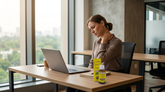 A woman in an office setting massages her neck in pain while a bottle of soothing oil sits on her desk next to her laptop.