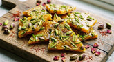 Close-up photograph of Persian pistachio Sohan brittle pieces, generously topped with pistachios, arranged on a rustic wooden cutting board with scattered dried rose petals and saffron threads.