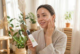 A beautiful young woman with radiant skin applying Botox Plus organic cream from a white tube, smiling in a bright, spa-like room with natural sunlight and green plants.